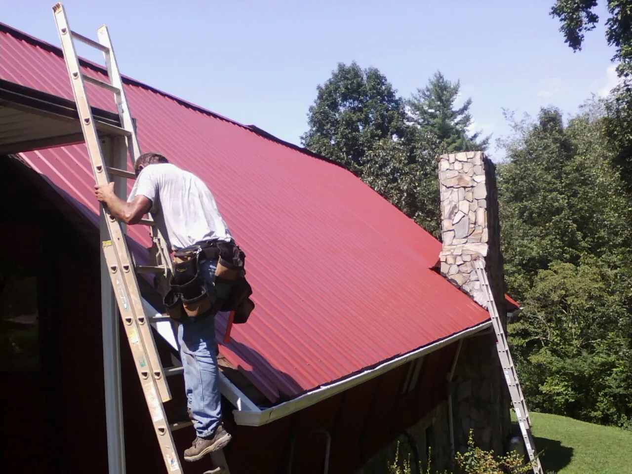 Roofer on a ladder beside a red metal roof and stone chimney — Soffit & Fascia Repair by Roofing Brothers in Elmer